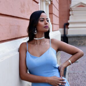 Stylish woman in a blue dress with jewelry poses elegantly against a brick wall outdoors.